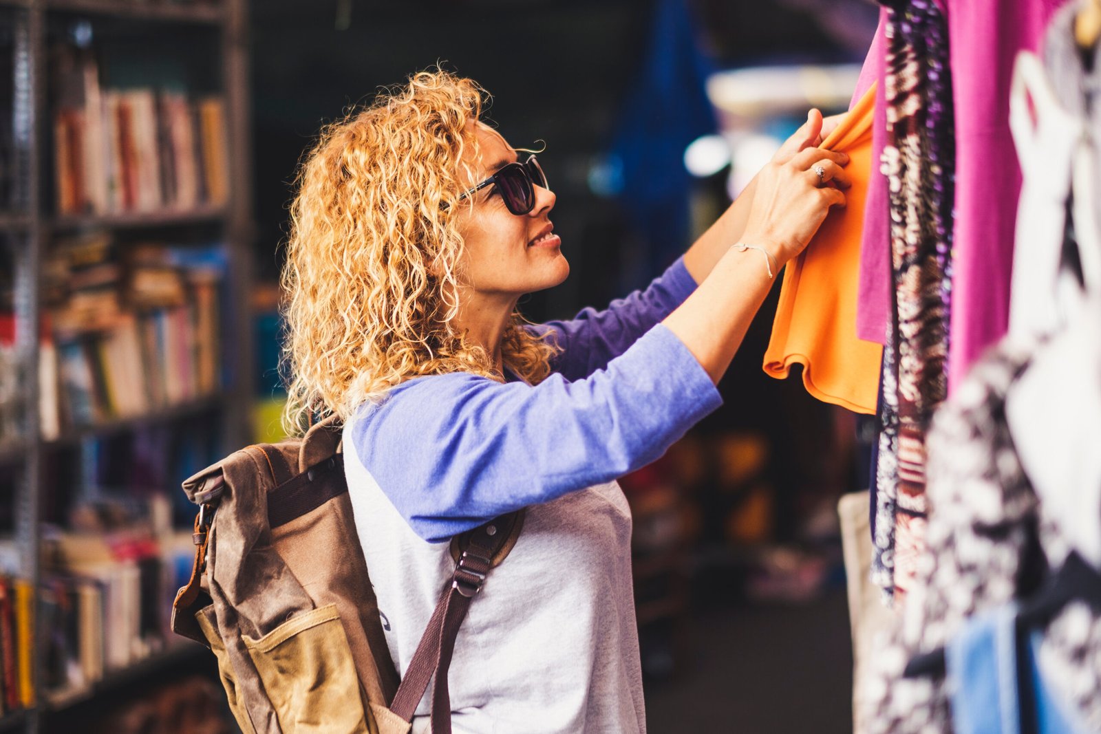 Cheerful traveler young blonde curly woman looking and choosing clothes at the used market during alternative vacation - shopping lifestyle concept for nice people